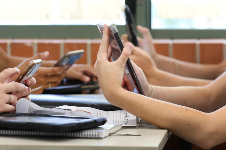 A table of high school students all holding multiple digital devices such as phones and tablets during a class lesson.