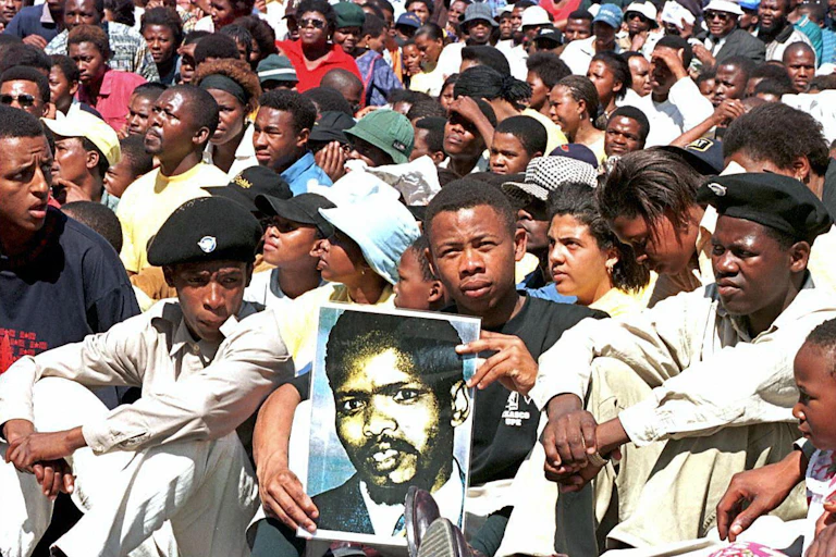 Crowd of seated people, with one holding up a poster of a man's face