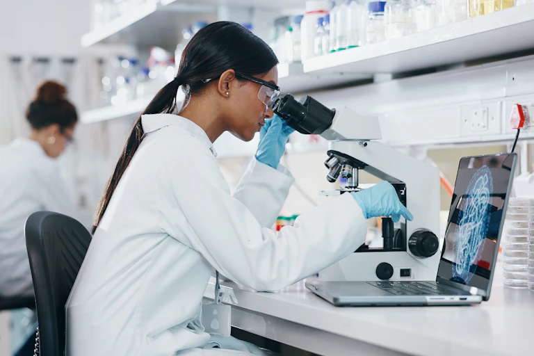 A woman in a lab coat with long dark hair looks into a microscope.