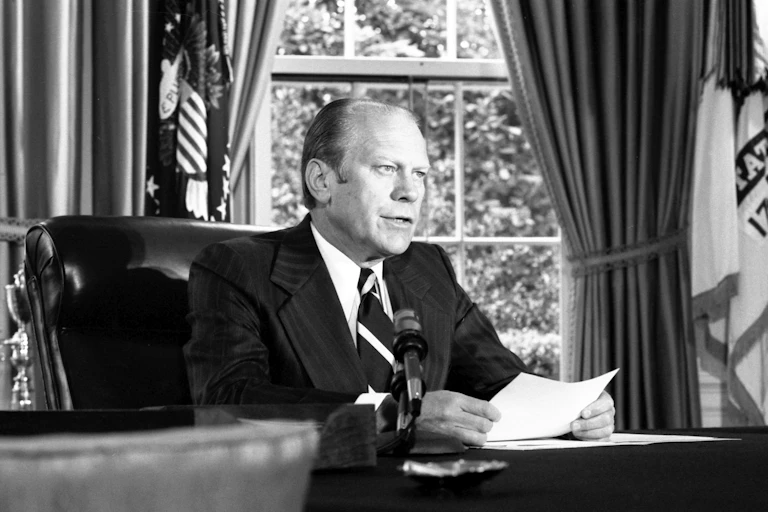 A man in a suit and tie sitting at a desk reading from a piece of paper in front of him.