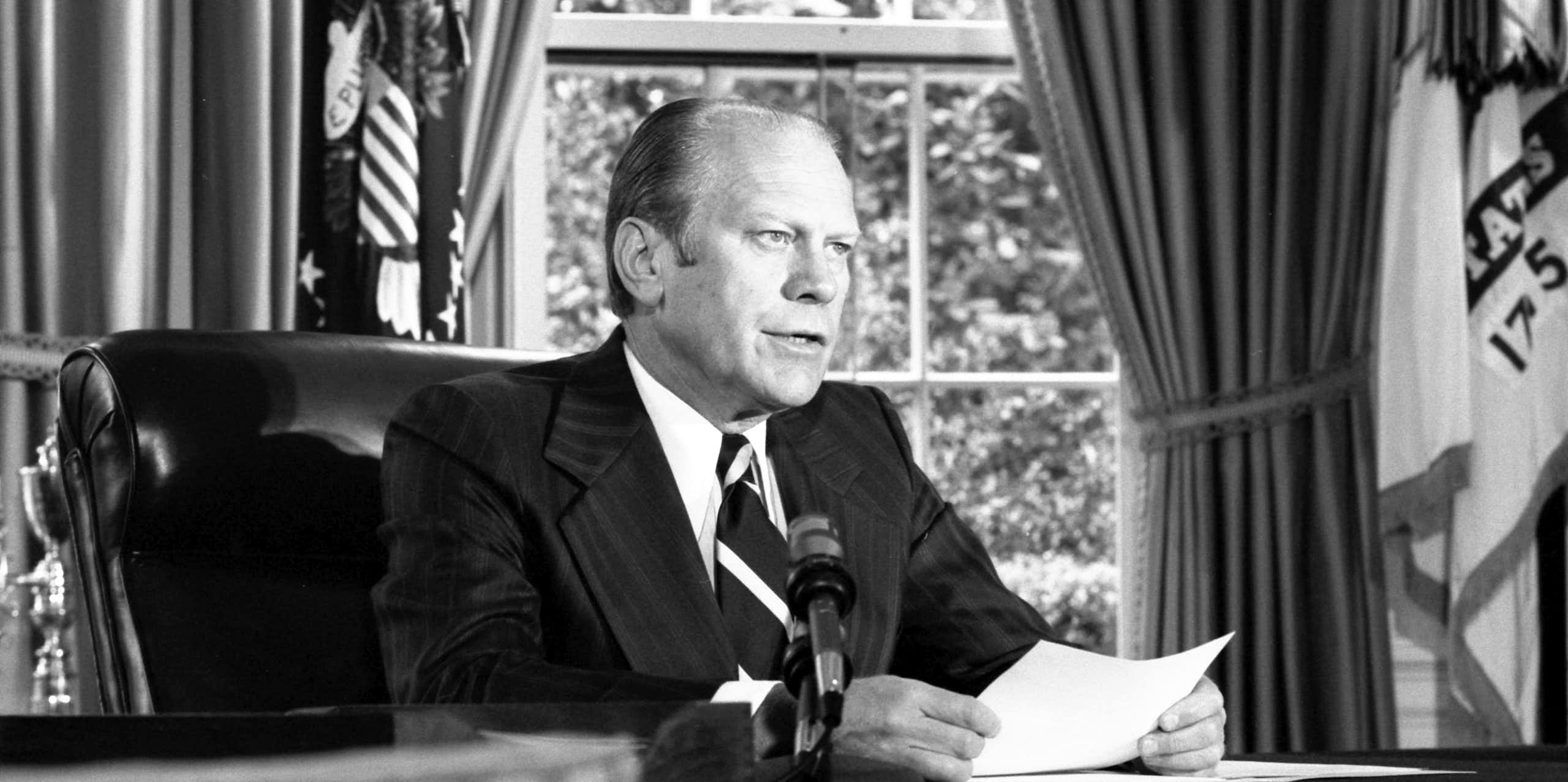 A man in a suit and tie sitting at a desk reading from a piece of paper in front of him.
