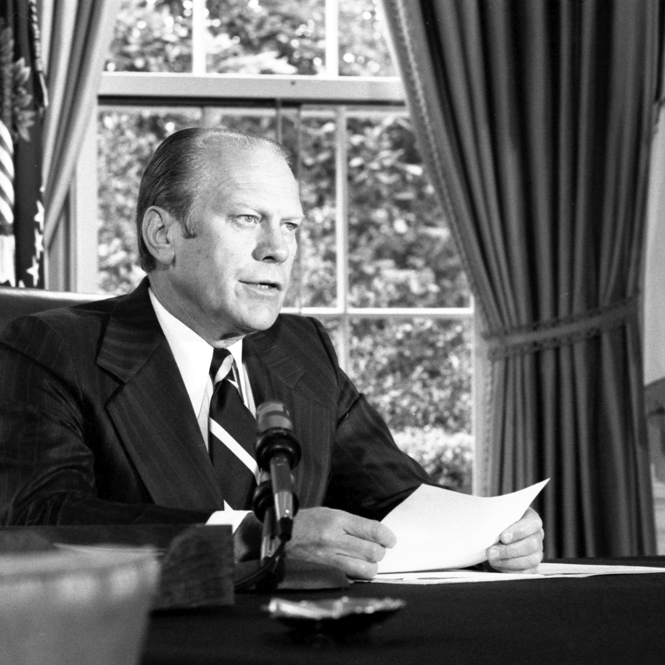 A man in a suit and tie sitting at a desk reading from a piece of paper in front of him.