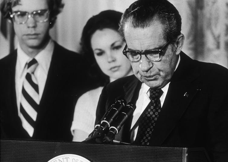 A dark-haired man in glasses, looking solemn, stands at a lectern and reads a speech into microphones.
