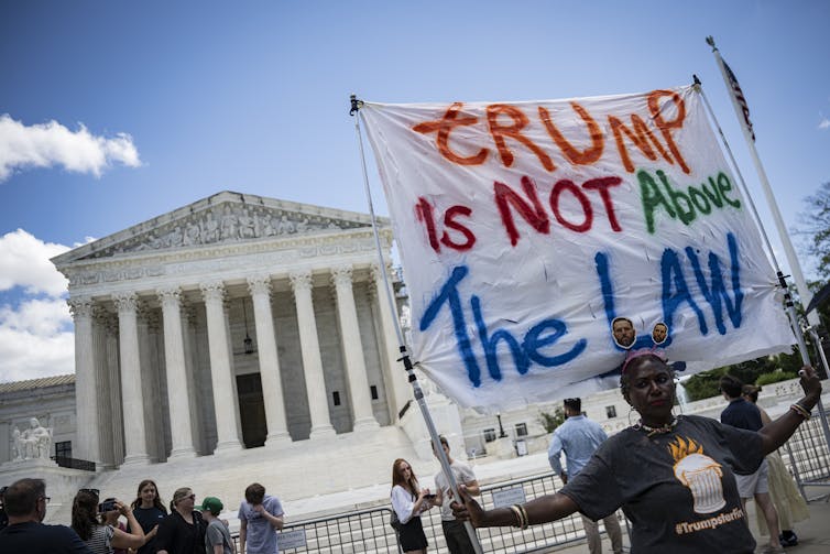 A person holding a banner that says 'Trump is NOT above the law' in front of a large while building.