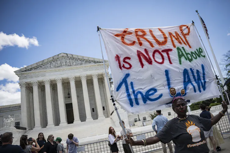 A person holding a banner that says 'Trump is NOT above the law' in front of a large while building.