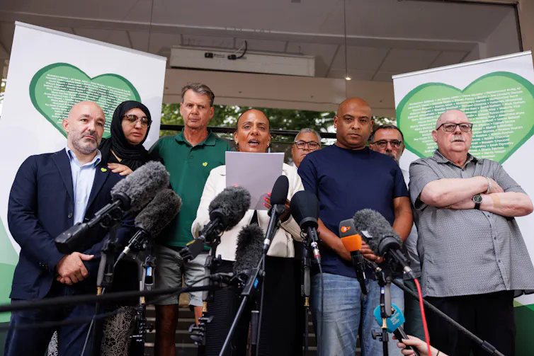 A racially diverse group of people standing together at a series of microphones, while a woman reads a statement