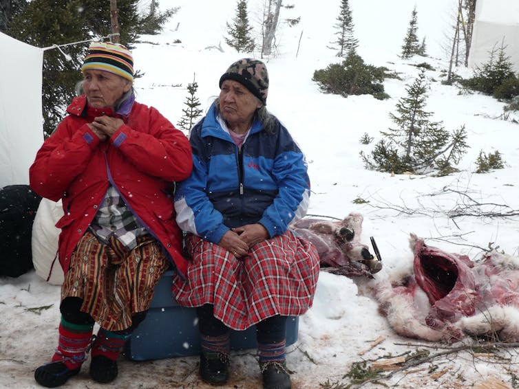 Two Innu elder women seated in a snowy landscape.