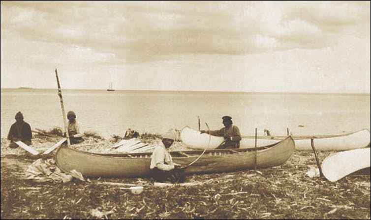 Innu people from 1920 making canoes on the shore of a lake.