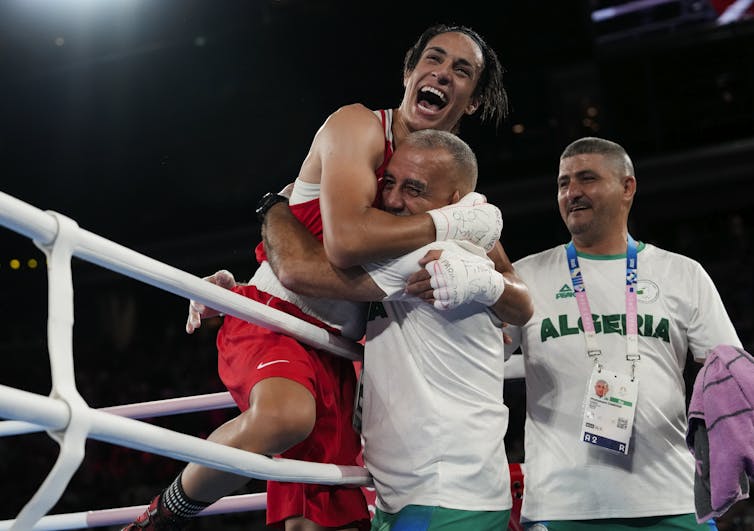 A smiling woman with boxing gloves on and red boxing shorts smiles joyously while being embraced by her coach.