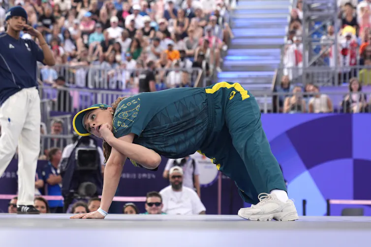 A woman in dark green breakdances in front of a crowd.