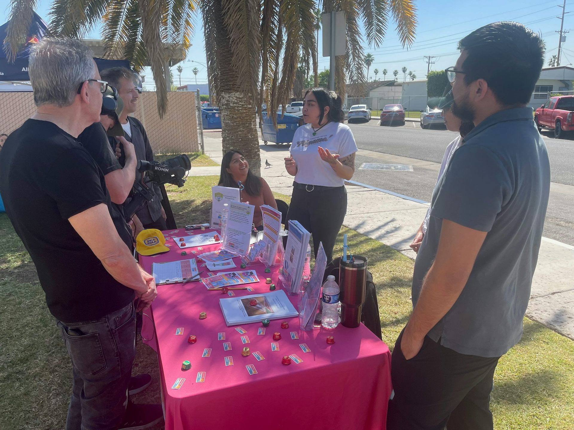 Young people stand at a table covered with brochures. They're talking with a videographer in the shade.