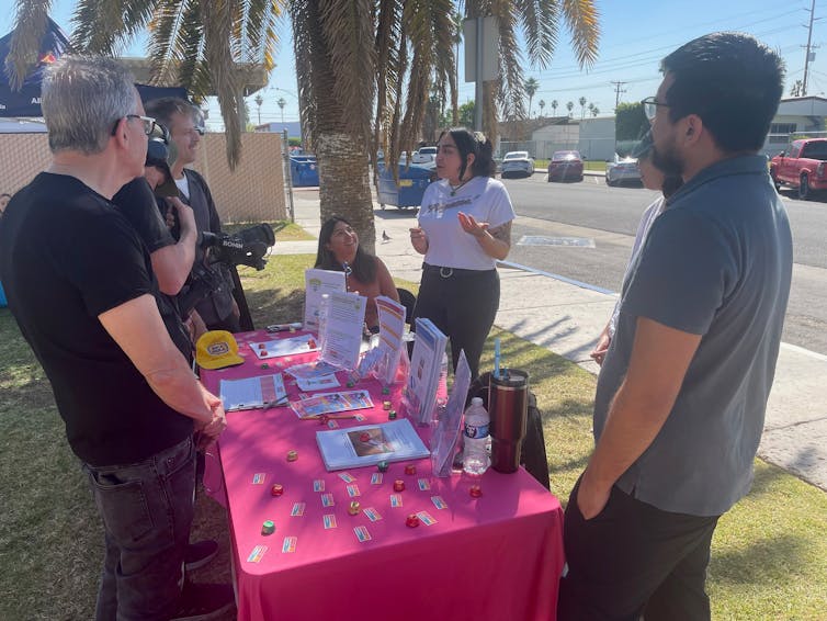 Young people stand at a table covered with brochures. They're talking with a videographer in the shade.