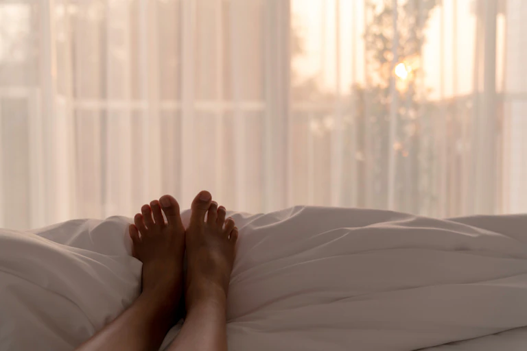 Person lying on bed, legs on top of covers, facing window, morning light shining through net curtains