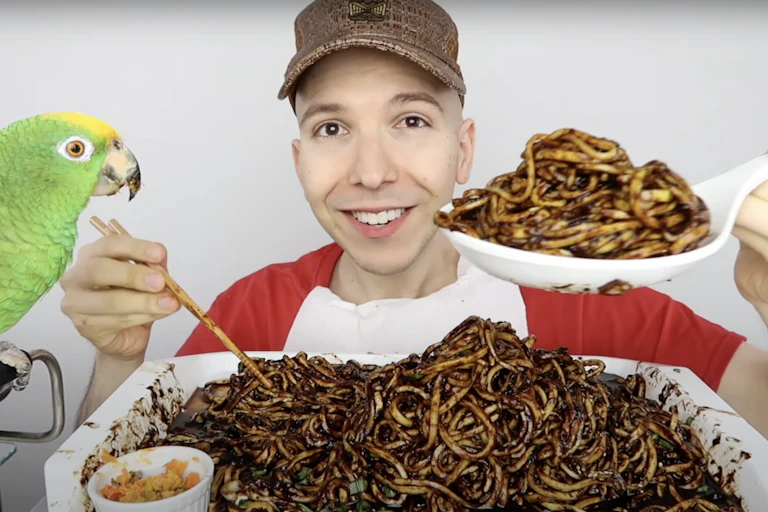 A YouTuber in a cap films a mukbang video with black bean noodles and a green parrot to his side