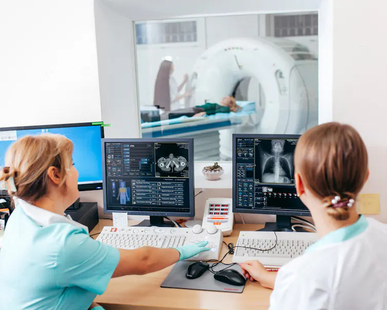 Deux infirmières regardent deux écrans d’ordinateur alors qu’un patient entre dans un scanner