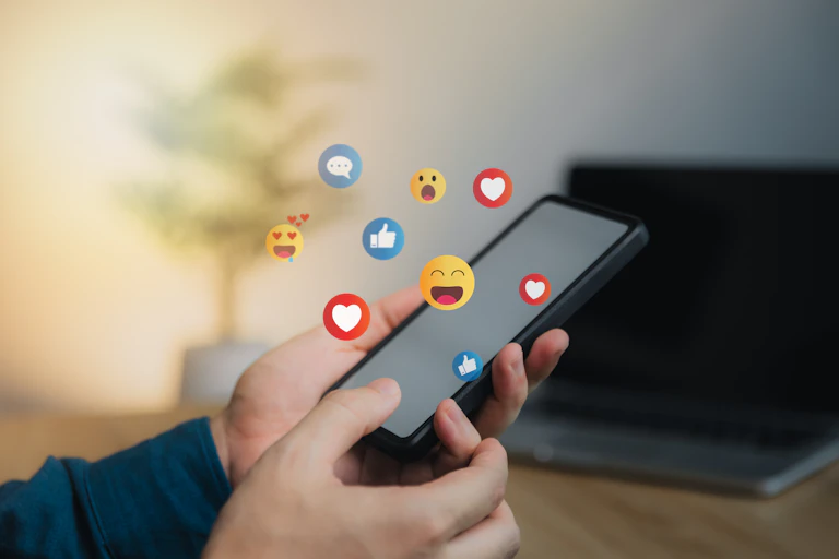 A young person holds a phone, with emojis, hearts and thumbs up images around it.