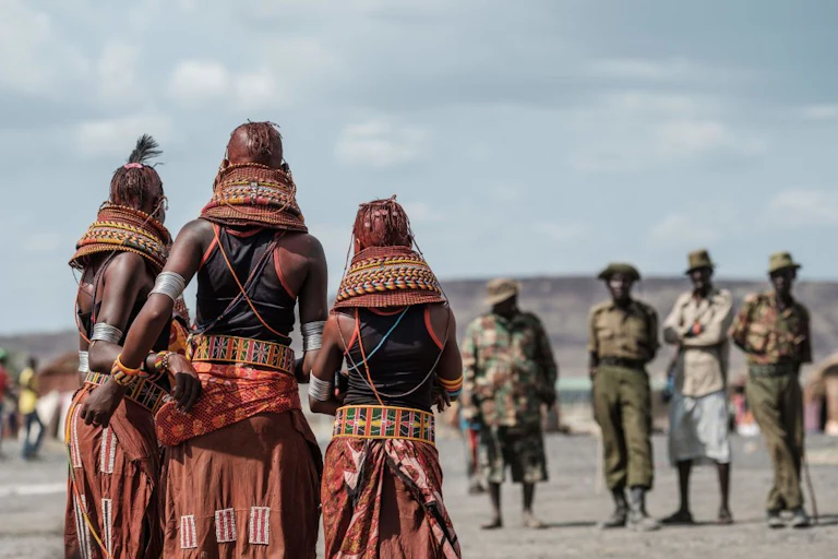 Three women in identical traditional dress and necklaces face a group of onlookers