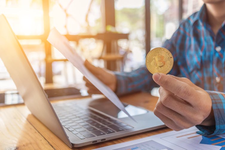 Man holds bitcoin in his hand as he works from laptop.