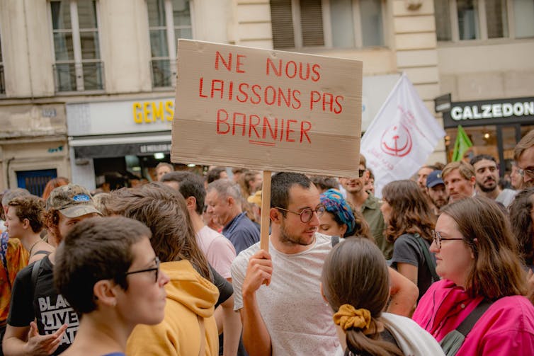 Um grupo de manifestantes com um homem no centro segurando uma placa que diz 'Ne nous laissons pas Barnier'