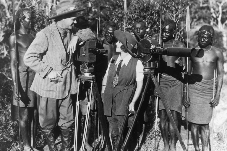 A vintage photograph of a European couple dressed in hats look at one another as they pose with two cameras in front of a row of shirtless African men holding spears, some of them bemused.