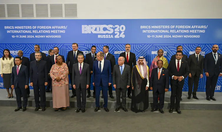 A group of men and women stand for a photo in front of a sign that reads BRICS 2024 Russia.