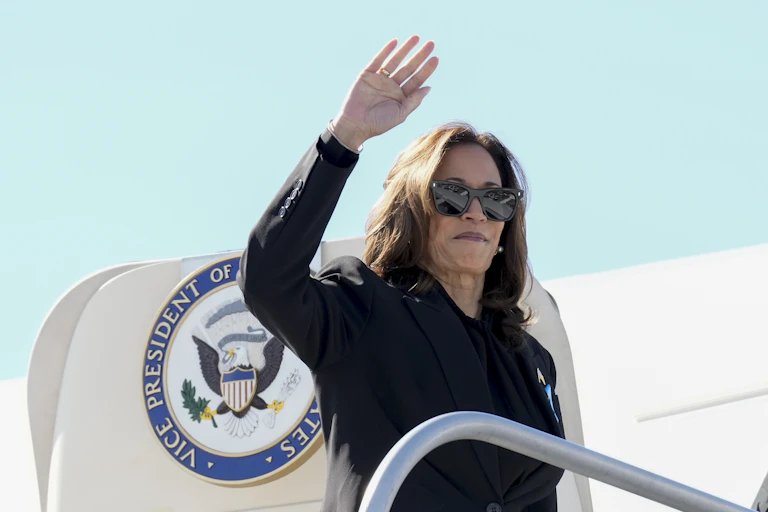 A dark-haired woman wearing sunglasses waves as she boards an aircraft on a sunny day.