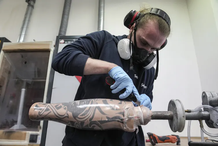 A man wearing a safety mask and gloves turns a prosthetic leg with tattoos on a lathe