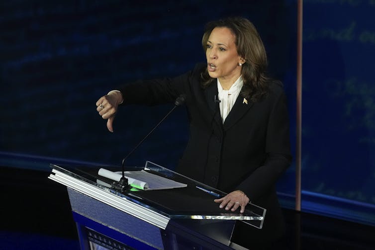 Kamala Harris stands behind a lecturn giving a thumbs down