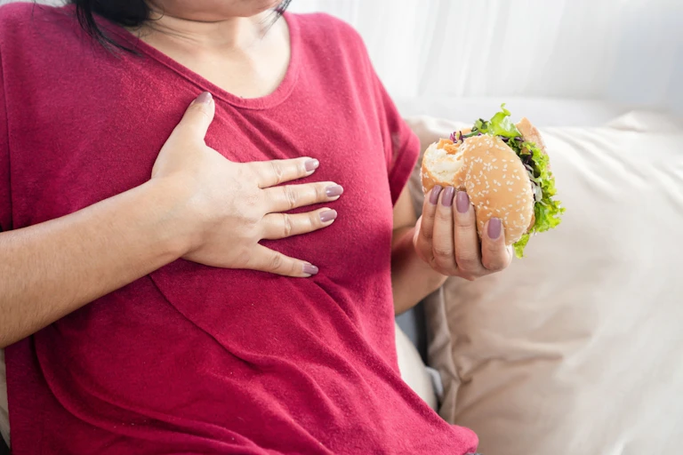 Woman sitting on sofa, one hand on chest, the other holding burger