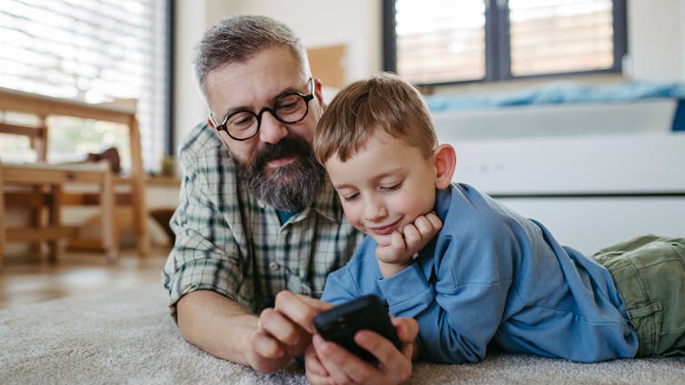 A man and a boy lie on the floor looking at a phone together.