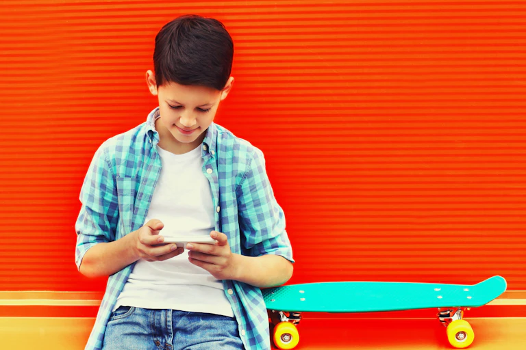 A teen boy against a bright red wall reads phone with a skateboard next to him.