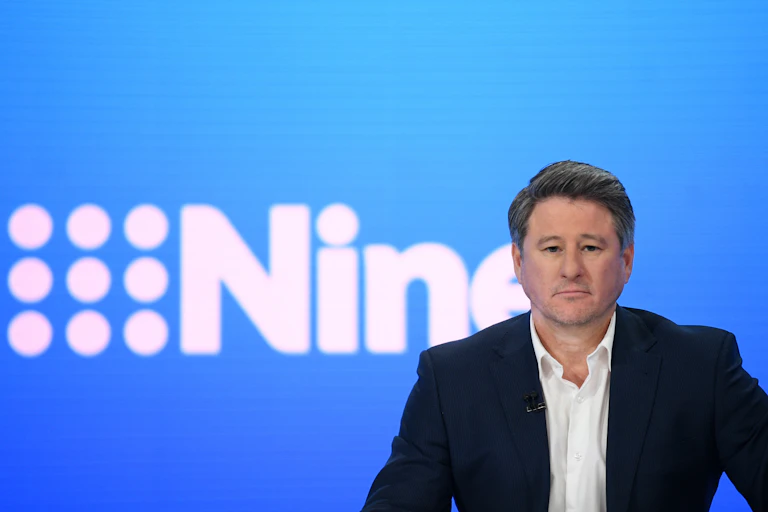 Man in black suit sits in front of Nine logo