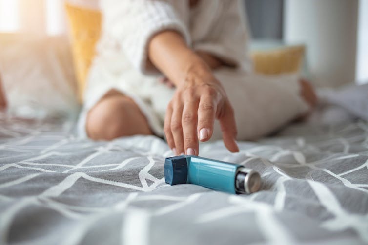 A hand reaches for an asthma inhaler on a bed.