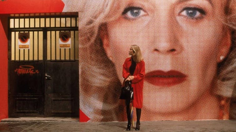 Una mujer con una gabardina roja espera delante de una pared con la foto de una mujer con labios rojos y fondo también rojo.