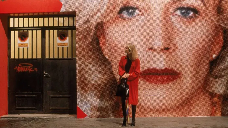Una mujer con una gabardina roja espera delante de una pared con la foto de una mujer con labios rojos y fondo también rojo.