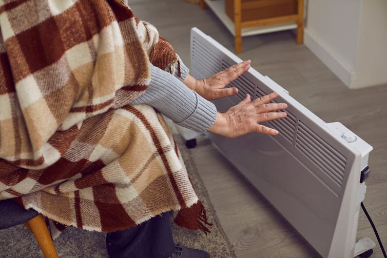 An old man covered in a blanket holds his hands to an electric space heater.