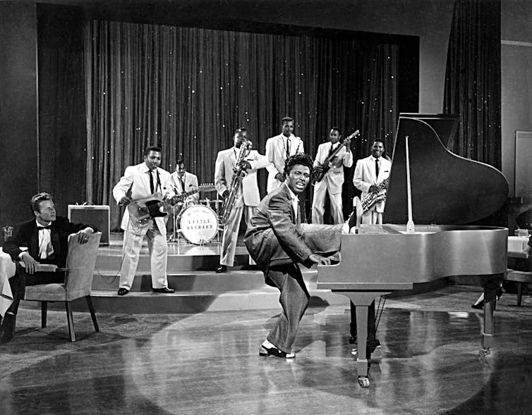 A black and white photo shows a young Little Richard playing the piano while standing on one leg (the other is improbably resting on the piano).