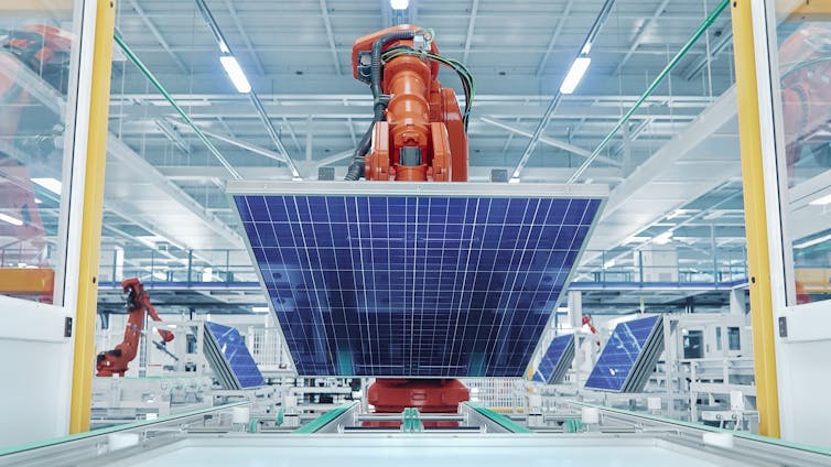 Huge orange robot arm lifts big new blue solar panel in brightly lit factory