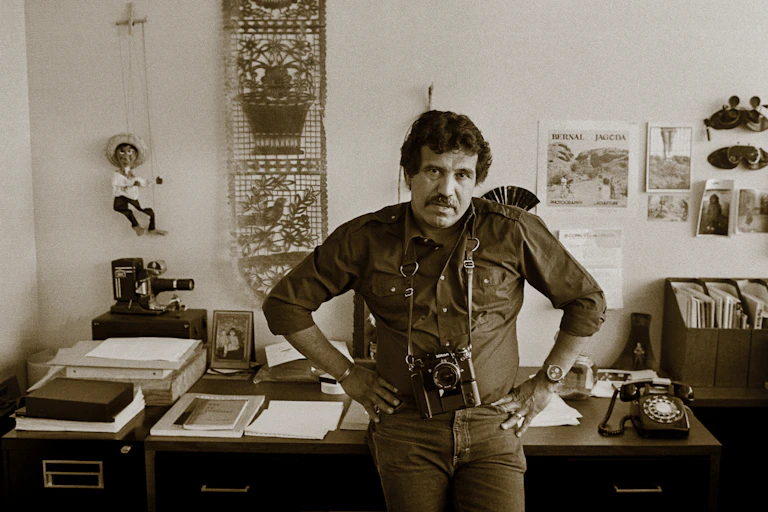 Sepia portrait of middle aged man standing in front of a desk with his hands on his hips and a camera hanging from his neck.