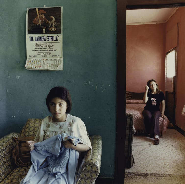 Photograph of a young woman sewing in the foreground, with another young woman sitting on a bed in the background.