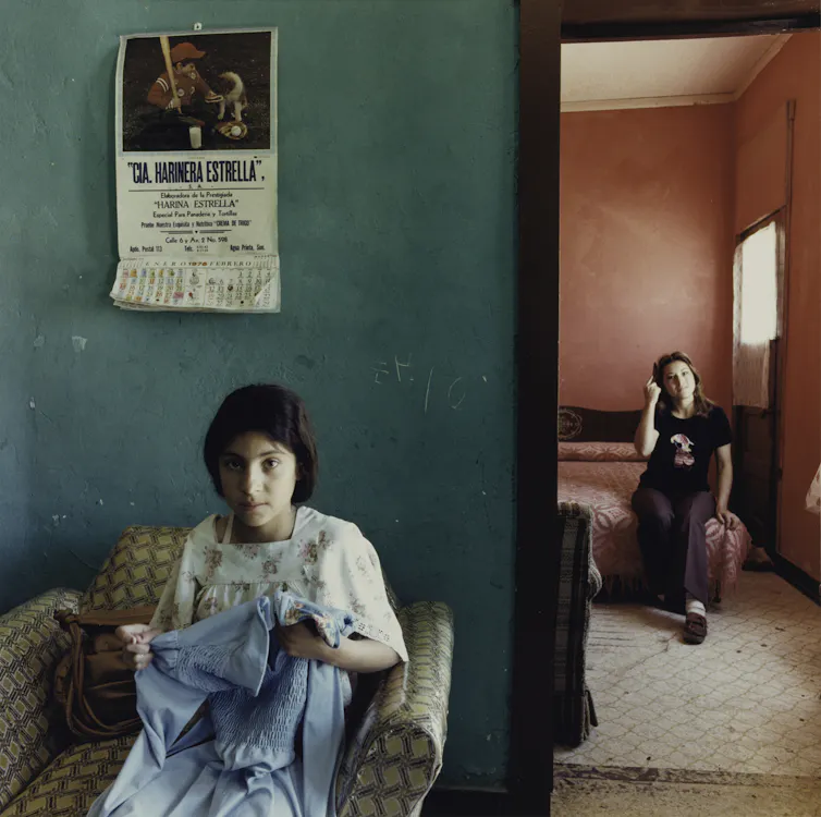 Photograph of a young woman sewing in the foreground, with another young woman sitting on a bed in the background.