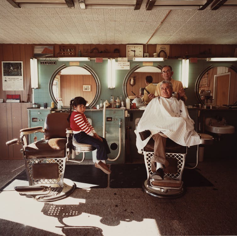 A little girl sits on a chair watching a barber cut the hair of his customer.