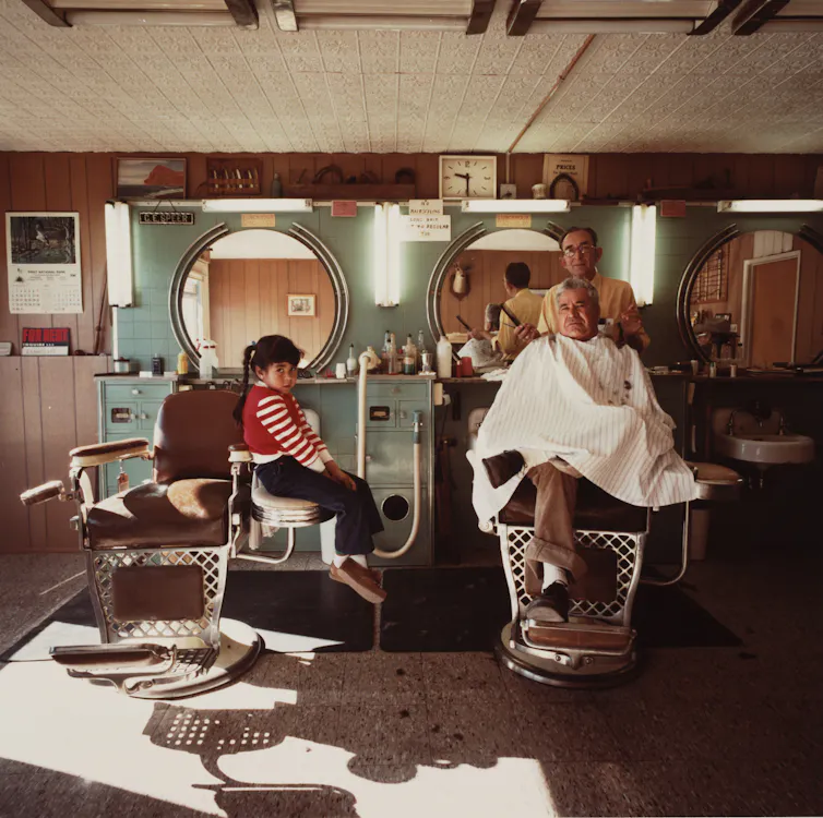 A little girl sits on a chair watching a barber cut the hair of his customer.