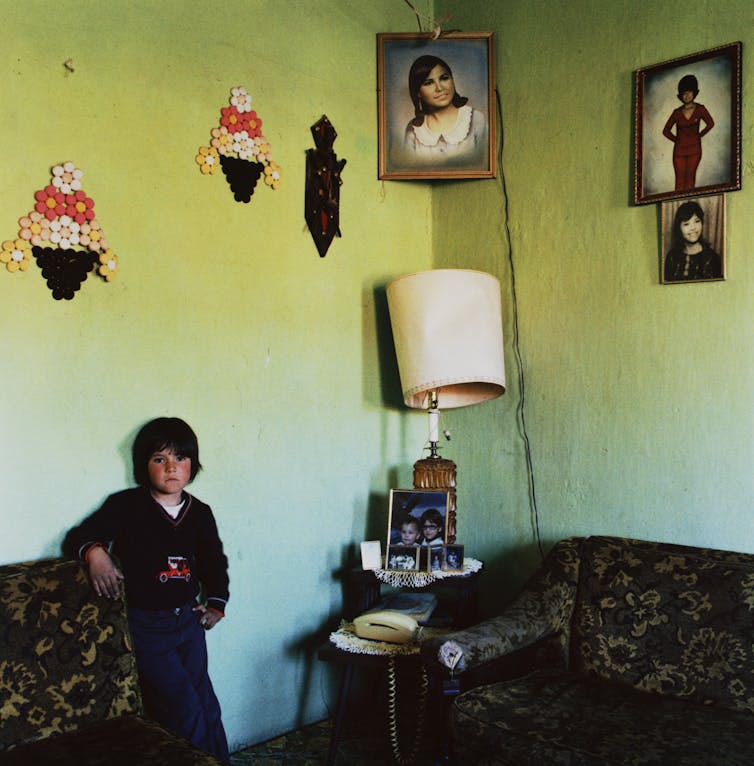 A little boy poses next to cushioned furniture in front of a green wall adorned with portraits and tchotchkes.