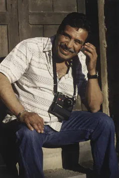 A middle-aged man with a mustache and wearing a camera around his neck smiles as he sits on concrete steps.