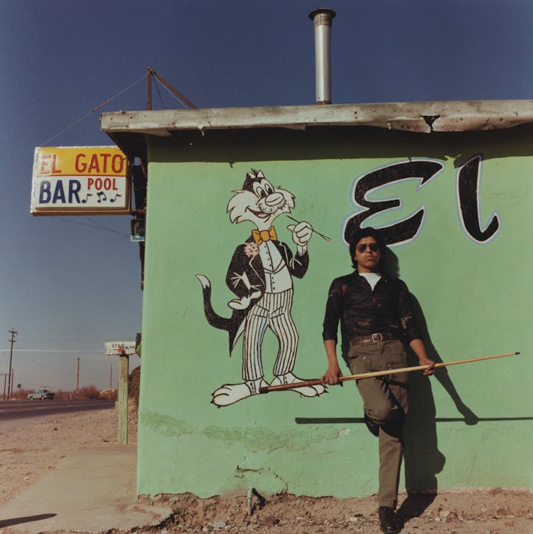 Young man wearing sunglasses holds a pool cue while posing against a green-walled bar called 'El Gato' in the desert.