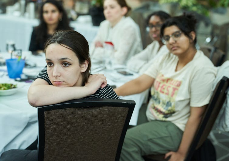 Young women sit around a table listening to someone speaking off-camera.