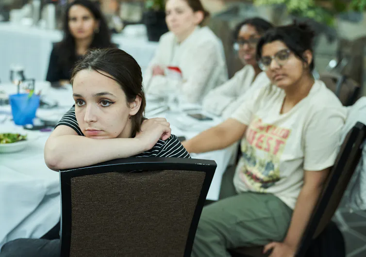 Young women sit around a table listening to someone speaking off-camera.