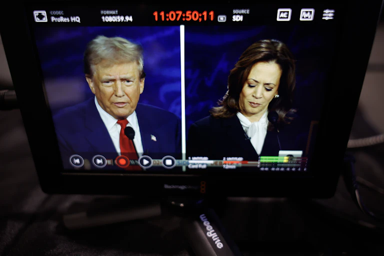 A man with a blue jacket and red tie is seen on a split screen with a woman with dark hair and a dark jacket looking down.