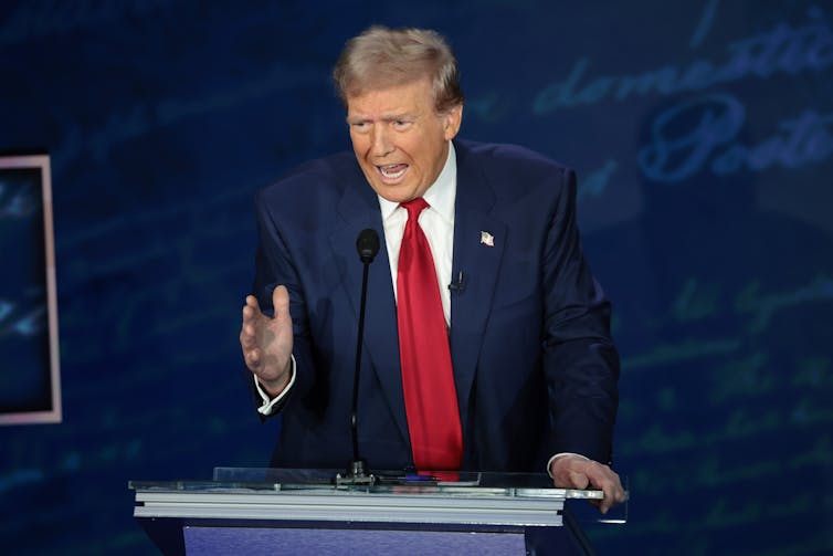 A man with a blue jacket and red tie stands at a podium and gestures with his hand.